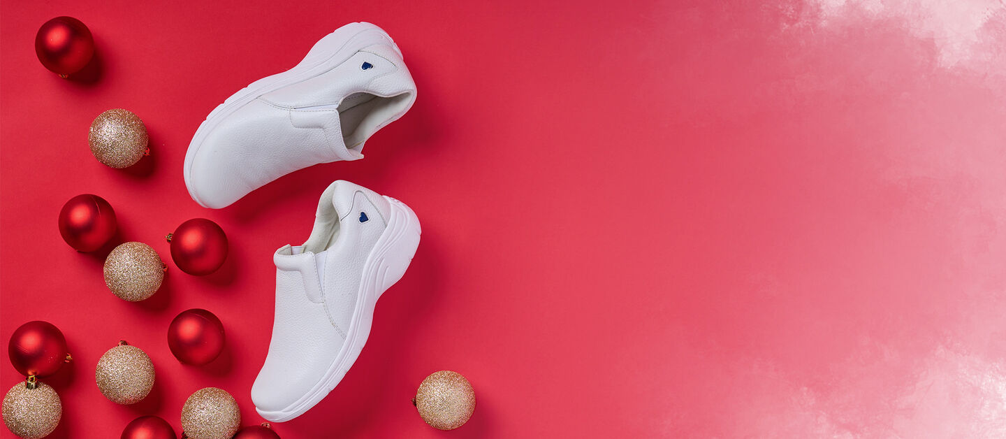 White Dove slip on shoes laid flat on a bright red background with Christmas bulbs decorating around the shoe. 